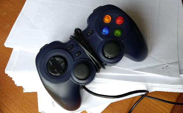 A video game controller lies on top of a stack of stapled papers on a wooden surface, the cord wrapped several times around its centre.